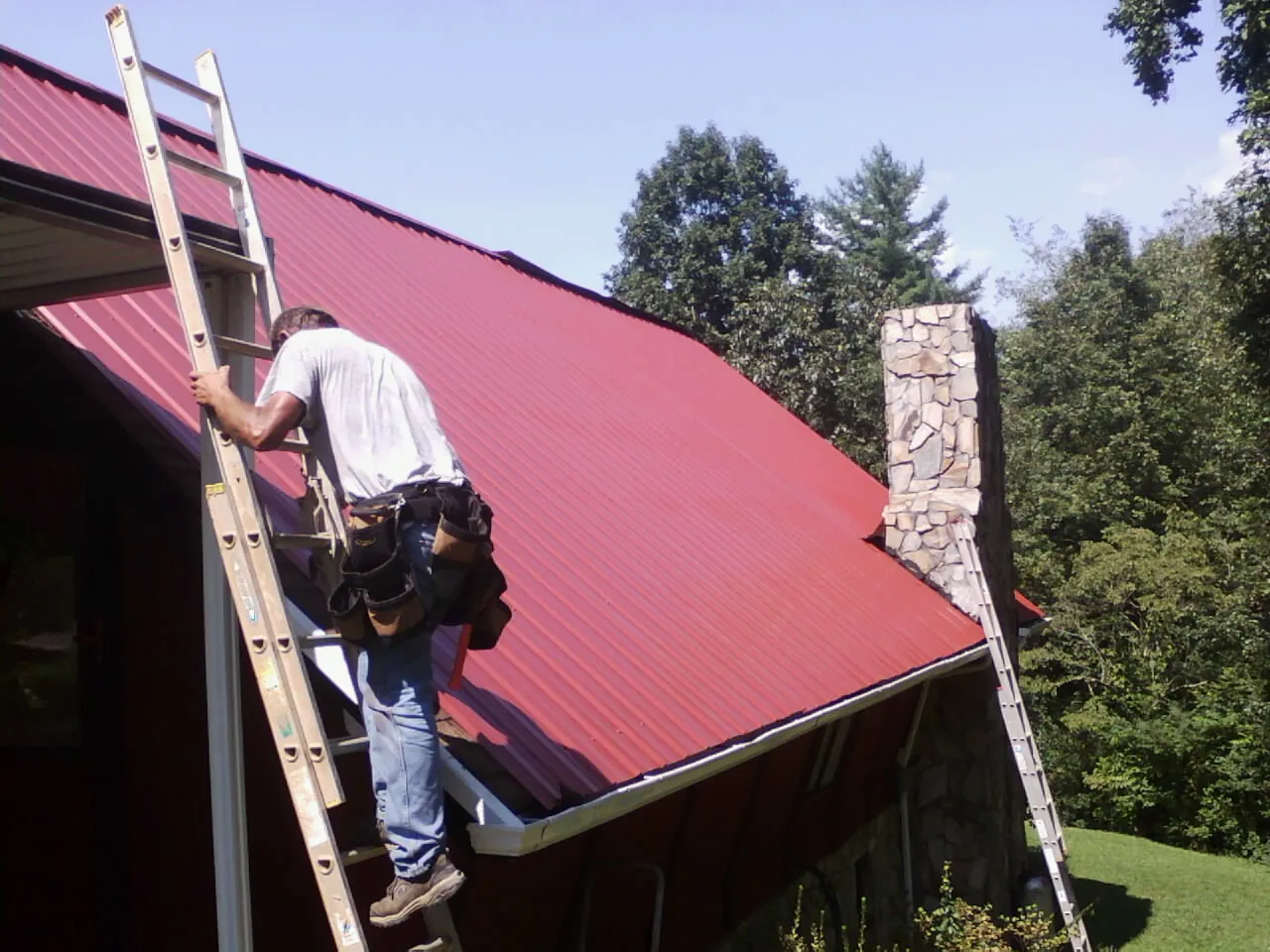 Roofer on a ladder beside a red metal roof and stone chimney — Soffit & Fascia Repair by Roofing Brothers in Kennebunkport